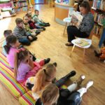 dzieci siedzą na kolorowym materacu, bibliotekarka czyta im książkę