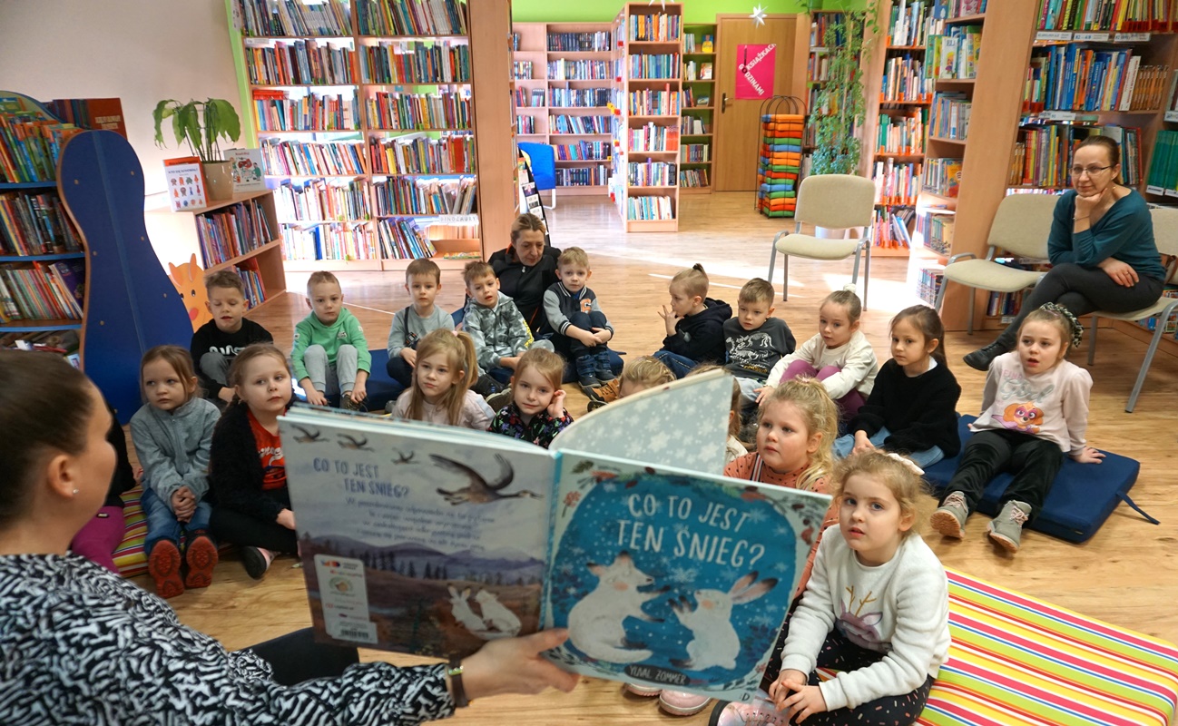 bibliotekarka pokazuje siedzącym przed nią dzieciom książkę którą aktualnie czyta