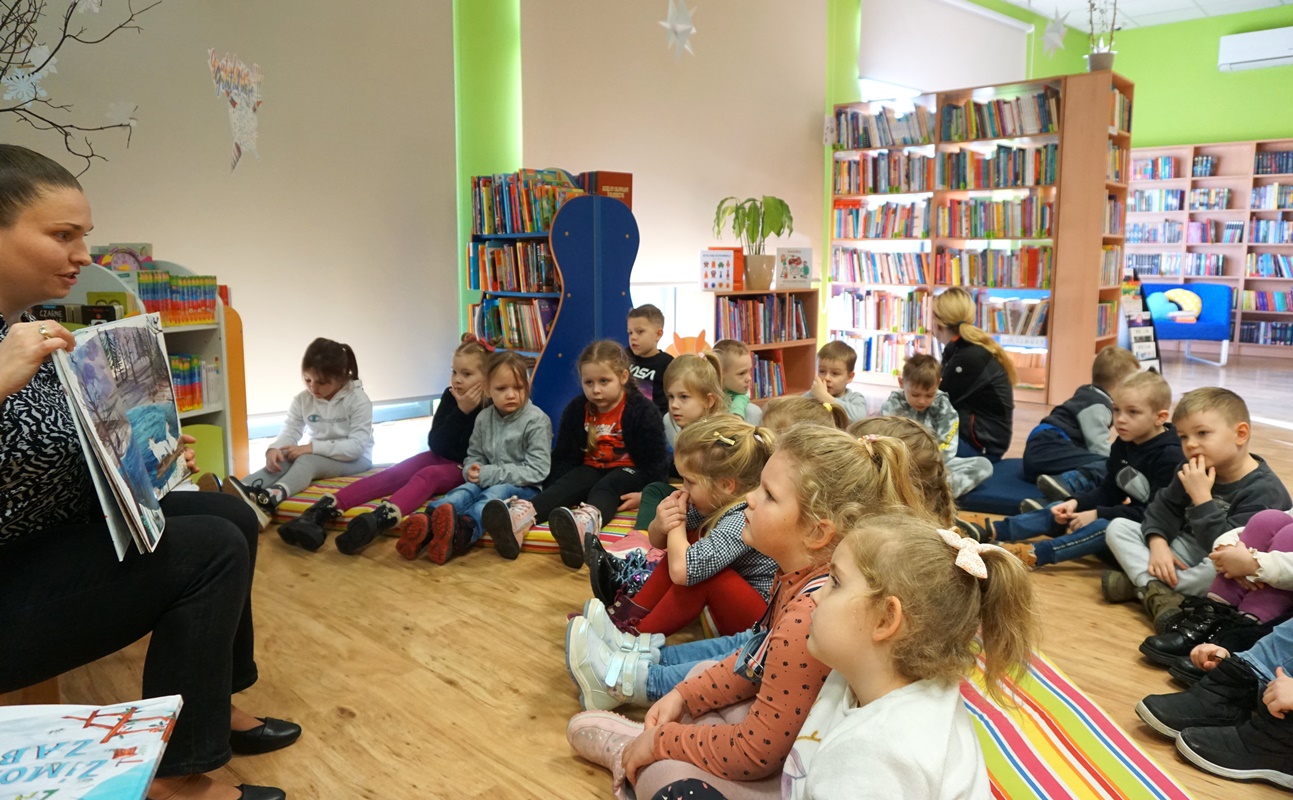 dzieci siedzące w półkolu słuchające czytanej książki