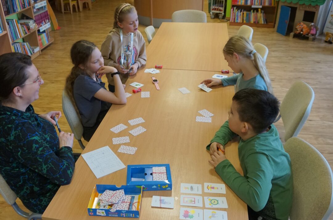 Czwórka dzieci i jedna dorosła kobieta siedzi przy stole i gra w grę w języku angielskim, jedna z dziewczynek przygląda się obrazkowi na karcie.