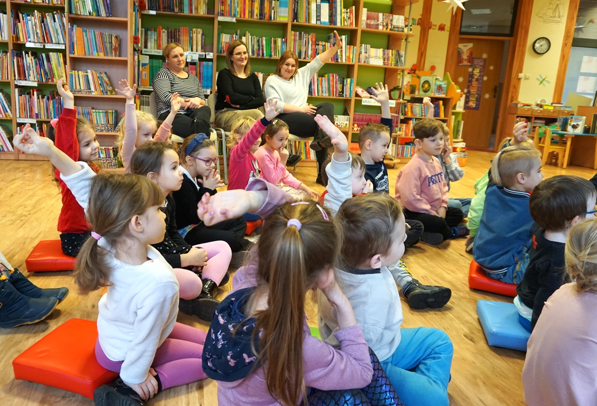 dzieci siedzą na kolorowych pufach, zgłaszają się chcąc odpowiedzieć na pytanie zadane przez prowadzącą zajęcia