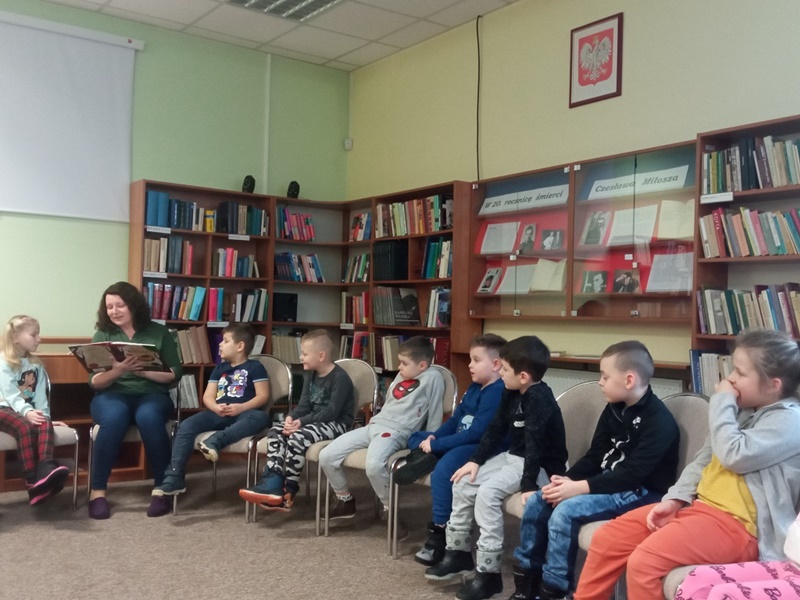 Dzieci biorące udział w zajęciach siedzą na krzesłach a bibliotekarka czyta im książkę