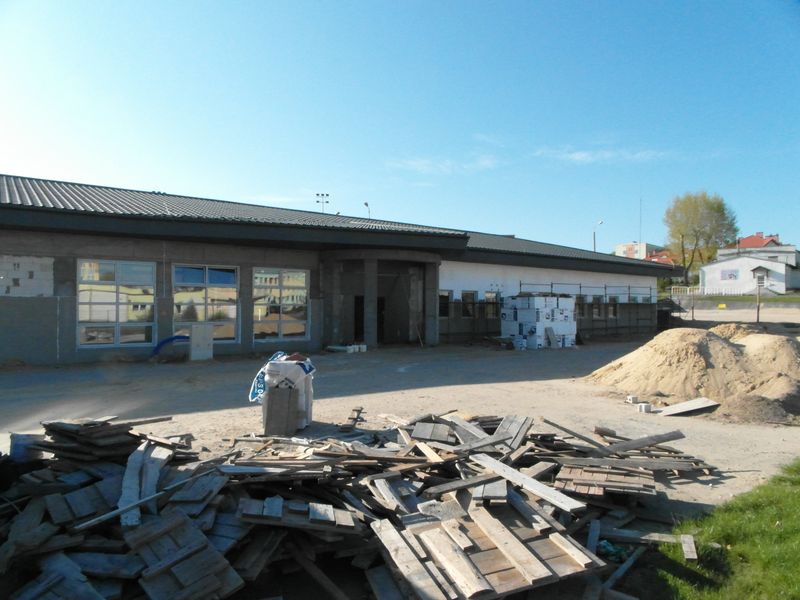 Budynek biblioteki podczas budowy, z przodu widać pozostałości desek oraz hałdę piasku, z tyłu nieotynkowany budynek o czarnym dachu z lachodachówki, przed budynkiem rusztowanie i częściowo ocieplona styropianem elewacja