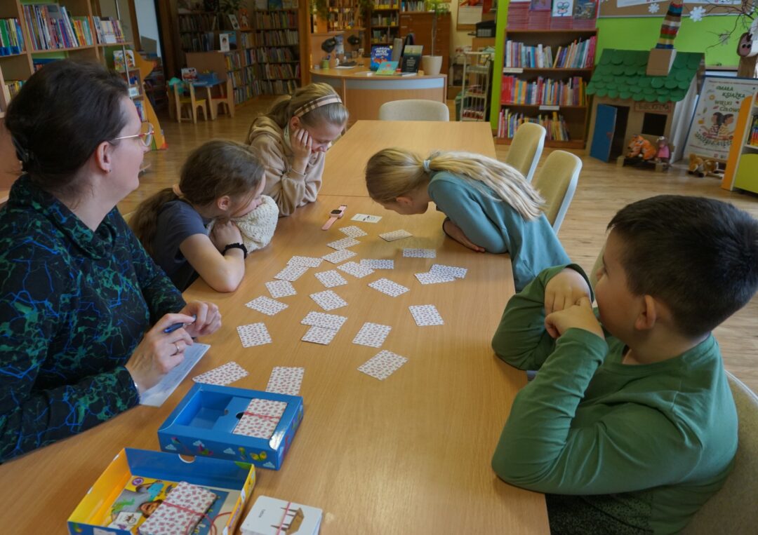 Uśmiechnięta grupa dzieci razem z prowadzącą zajęcia siedzi przy stole gra w grę z rodzaju memory dotyczącą angielskich słówek, wszyscy się uśmiechają.