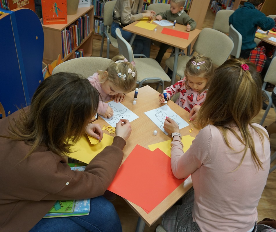 dwie dziewczynki smarują klejem wydrukowany obrys kubusia puchatka i przyklejają kolorowe kawałki papieru, dwie opiekunki siedzące razem z nimi przyglądają się ich pracy