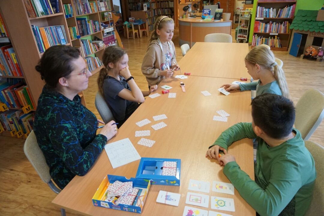 Trzy dziewczynki, jeden chłopiec i bibliotekarka korzystają z kolorowych kart przedstawiających przedmioty i osoby, wszystkie obrazki podpisane są w języku angielskim