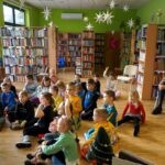 dzieci siedzą przed bibliotekarką któa czyta książkę, za dziećmi na krzesłach siedzą opiekunki obserwujące grupę