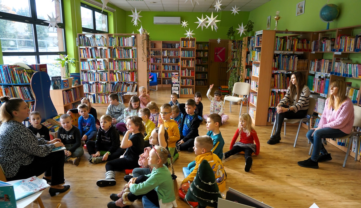dzieci siedzą przed bibliotekarką któa czyta książkę, za dziećmi na krzesłach siedzą opiekunki obserwujące grupę