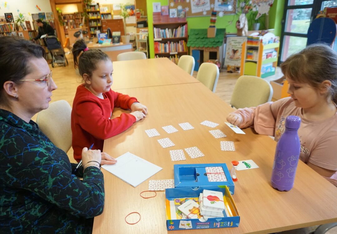 Dwie dziewczynki, razem z bibliotekarką prowadzącą zajęcia siedzą przy stole i korzystając z kolorowych kart uczą cię słówek w języku angielskim.