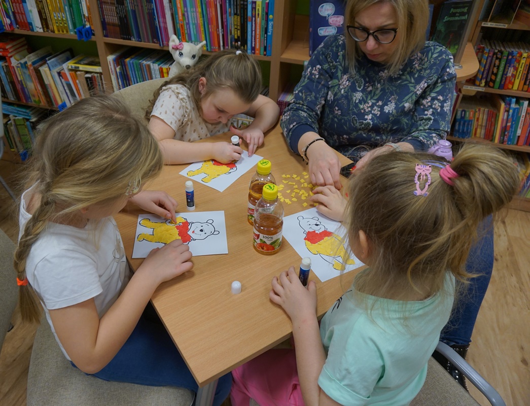 trójka dzieci pracująca nad swoimi obrazkami, opiekunka siedząca z nimi pomaga jednej z dziewczynek przyklejać żółte skrawki papieru