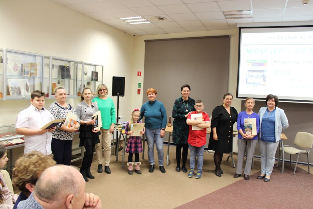 Grupowe zdjęcie uczestników i uczestniczek konkursów wraz z pracownikami biblioteki, widać 11 osób w tym dwoje dzieci, grupa stoi w półkolu, każda osoba trzyma wręczone dyplomy i nagrody książkowe.