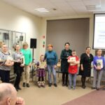 Grupowe zdjęcie uczestników i uczestniczek konkursów wraz z pracownikami biblioteki, widać 11 osób w tym troje dzieci, grupa stoi w półkolu, każda osoba trzyma wręczone dyplomy i nagrody książkowe.