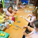 5 dziewczynek uczestniczących w zajęciach siedzi przy stołach i zaplata kolorowe sznurki według instrukcji otrzymanych od bibliotekarki prowadzącej zajęcia