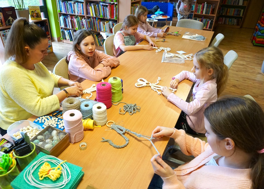 5 dziewczynek uczestniczących w zajęciach siedzi przy stołach i zaplata kolorowe sznurki według instrukcji otrzymanych od bibliotekarki prowadzącej zajęcia