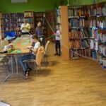 Uczestniczki zajęć razem z bibliotekarką prowadzącą zajęcia, stoją w kilkumetrowych odstępach od siebie i trzymają między sobą sznurki do makramy