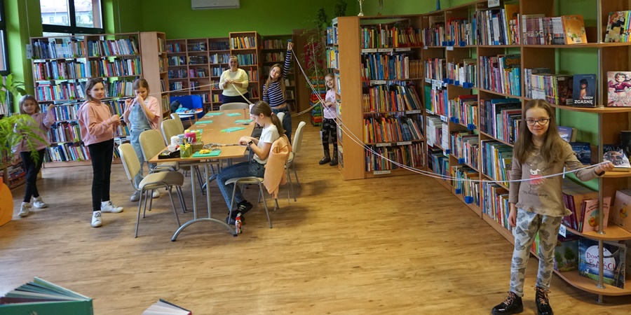 Uczestniczki zajęć razem z bibliotekarką prowadzącą zajęcia, stoją w kilkumetrowych odstępach od siebie i trzymają między sobą sznurki do makramy