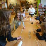 Dwie dziewczynki stoją przy stole i mają na blacie rozciągnięty sznurek, pięć innych siedzi na krzesłach i słucha instrukcji wydawanych przez bibliotekarkę prowadzącą zajęcia