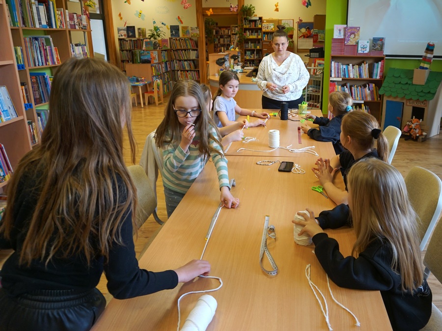 Dwie dziewczynki stoją przy stole i mają na blacie rozciągnięty sznurek, pięć innych siedzi na krzesłach i słucha instrukcji wydawanych przez bibliotekarkę prowadzącą zajęcia