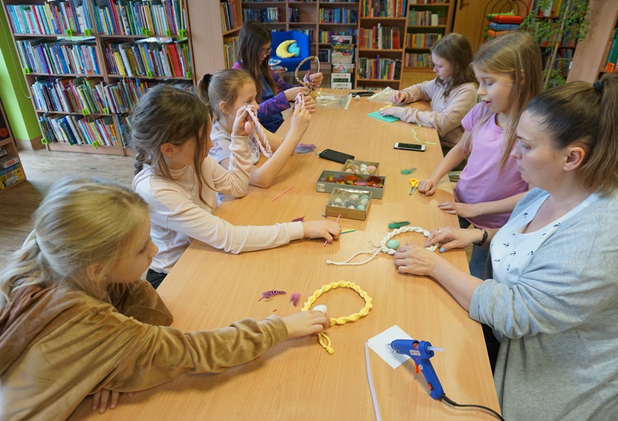 Uczestniczki zajęć pod okiem bibliotekarki formują zaplecione sznurki na kształt koła