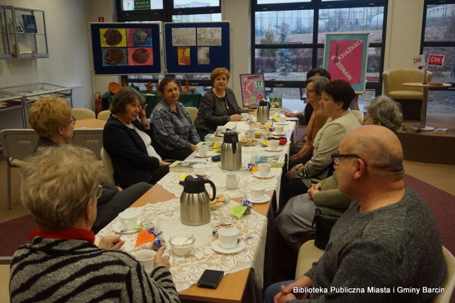 Uczestniczki i uczestnik spotkania siedzą wzdłuż stołów i rozmawiają o przeczytanej książce, na stołach znajduje się poczęstunek w formie kawy, herbaty i ciastek