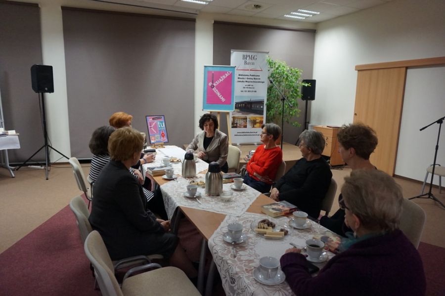 Osiem uczestniczek spotkania siedzi na krzesłach wzdłuż stołów, na których znajdują się książki, filiżanki z kawą i herbatą, oraz ciastka na talerzykach
