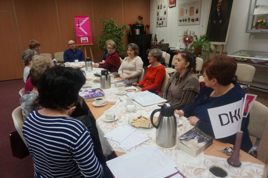 uczestniczki i uczestnik dyskusyjnego klubu książki siedzą przy stołach ustawionych w rzędzie, na stołach położone są koronkowe obruski, stoją ciastka, termosy z kawą i herbatą, leżą książki, w wazoniku na stole włożone są chorągiewki z logo DKK