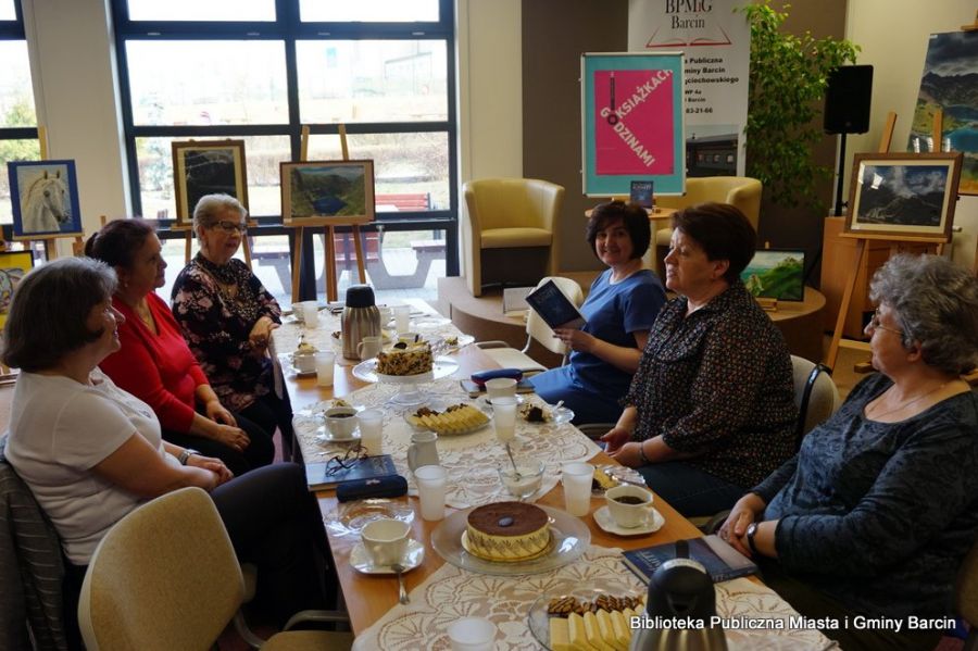 Uczestniczki spotkania siedzą wzdłuż stołów na których znajduje sie poczęstunek w formie kawy, herbaty, ciastek i tortu, rozmawia między sobą na temat przeczytanej ksiażki