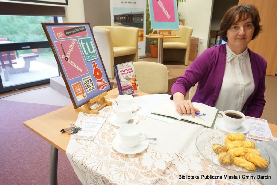 Bibliotekarka prowadząca klub DKK siedzi przy stole, po jej prawej stronie stoi plakat nawiązujący do działalności klubów DKK