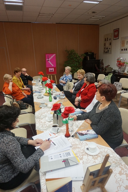 Uczestnicy spotkania siedzą zebrani wokół stołów ustawionych w rzędzie, na stole znajduje się poczęstunek w formie kawy, herbaty i ciastek, stół ozdabiają białe serwetki i wazoniki z papierowymi różami, prowadząca spotkanie robi notatki do sprawozdania.