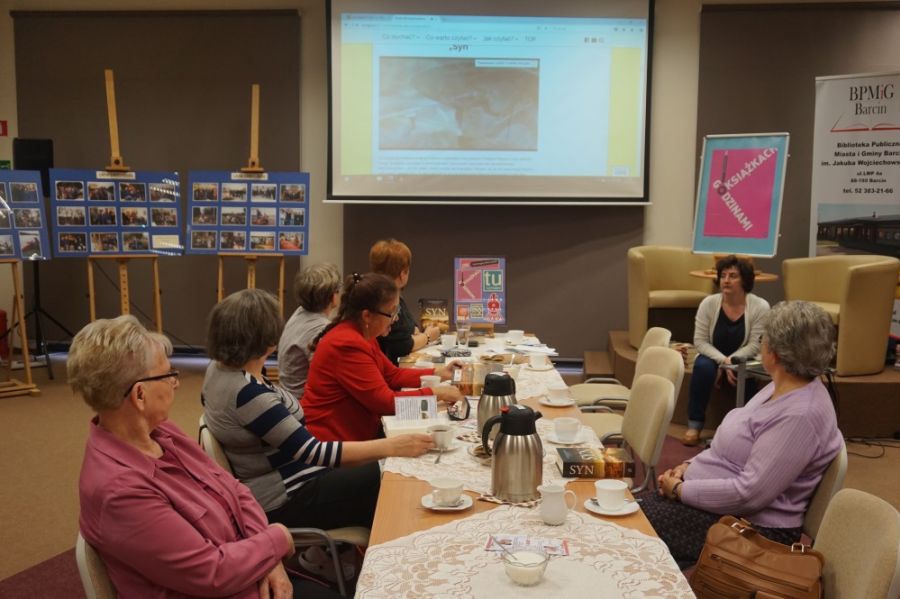 6 uczestniczek siedzi przy stole i patrzy na ekran projektora, siódma osoba siedzi przed komputerem w niedużej odległości od nich, szykuje się do włączenia filmu związanego z książką