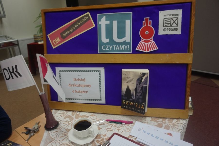 Tablica z niebieskim tle powieszone na niej jest logo DKK, grafika przypominająca lokomotywę, logo Instytuty Książki, kartka z napisem "Dzisiaj dyskutujemy o książce", oparta jest również omawiana książka