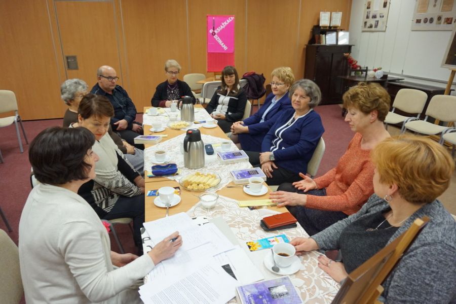 9 uczestniczek i 1 uczestnik Klubu siedzą przy stołach ustawionych w rzędzie i rozmawiają o przeczytanej książce, na stołach widać kawę i herbatę w termosach, ciastka na talerzykach a przed uczestnikami leżą omawiane ksiażki