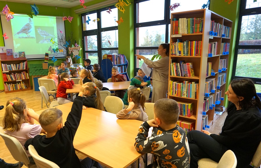 dzieci siedzą przy stolikach, jeden chłopiec podnosi rękę, bibliotekarka wskazuje na kogoś palcem