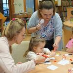 Bibliotekarka prowadząca zajęcia pokazuje dziewczynce gdzie ma przykleić posmarowane klejem białe kółko