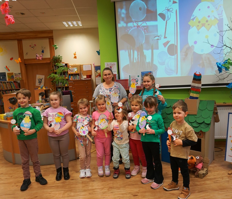 Grupowe zdjęcie uczestników zajęć, widać 9 dzieci i bibliotekarkę prowadzącą zajęcia, wszyscy z uśmiechami na twarzach pokazują prace wykonane podczas zajęć.