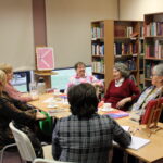 Uczestniczki Dyskusyjnego Klubu Książki zebrane wokół stołów na których leżą książki, stoją filiżanki z kawą lub herbatą z uśmiechami rozmawiają między sobą o omawianej książce, w tle stoi plakat DKK (różowe tło z tekstem Godzinami o Książkach)