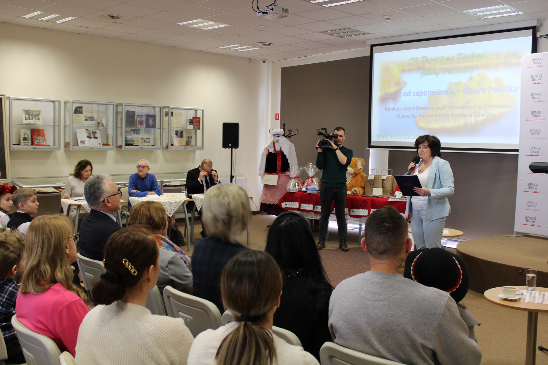 Dyrektorka biblioteki wita zebraną publiczność, kamerzysta z Telewizji Lokalnej nagrywa spotkanie