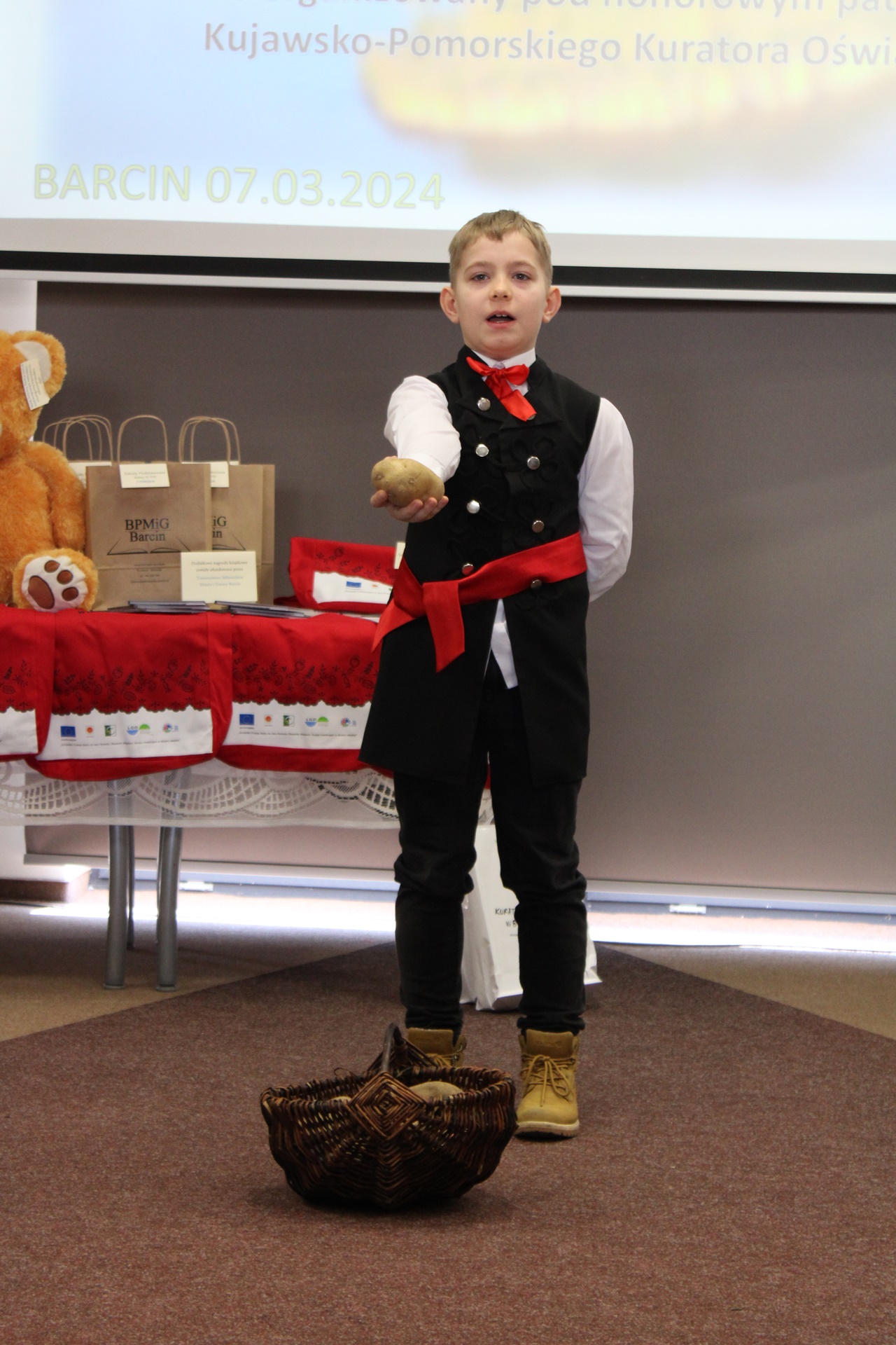 Chłopiec w ludowym stroju trzyma w ręku ziemniaka, przed nim w koszyku leżą inne ziemniaki, chłopiec recytuje wiersz konkursowy.