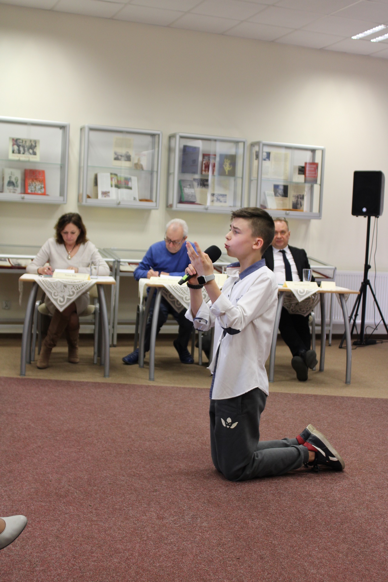 Chłopak w białej koszuli siwych spodniach i adidasach, podczas występu klęczy na wykładzinie i gestykulując recytuje wiersz.