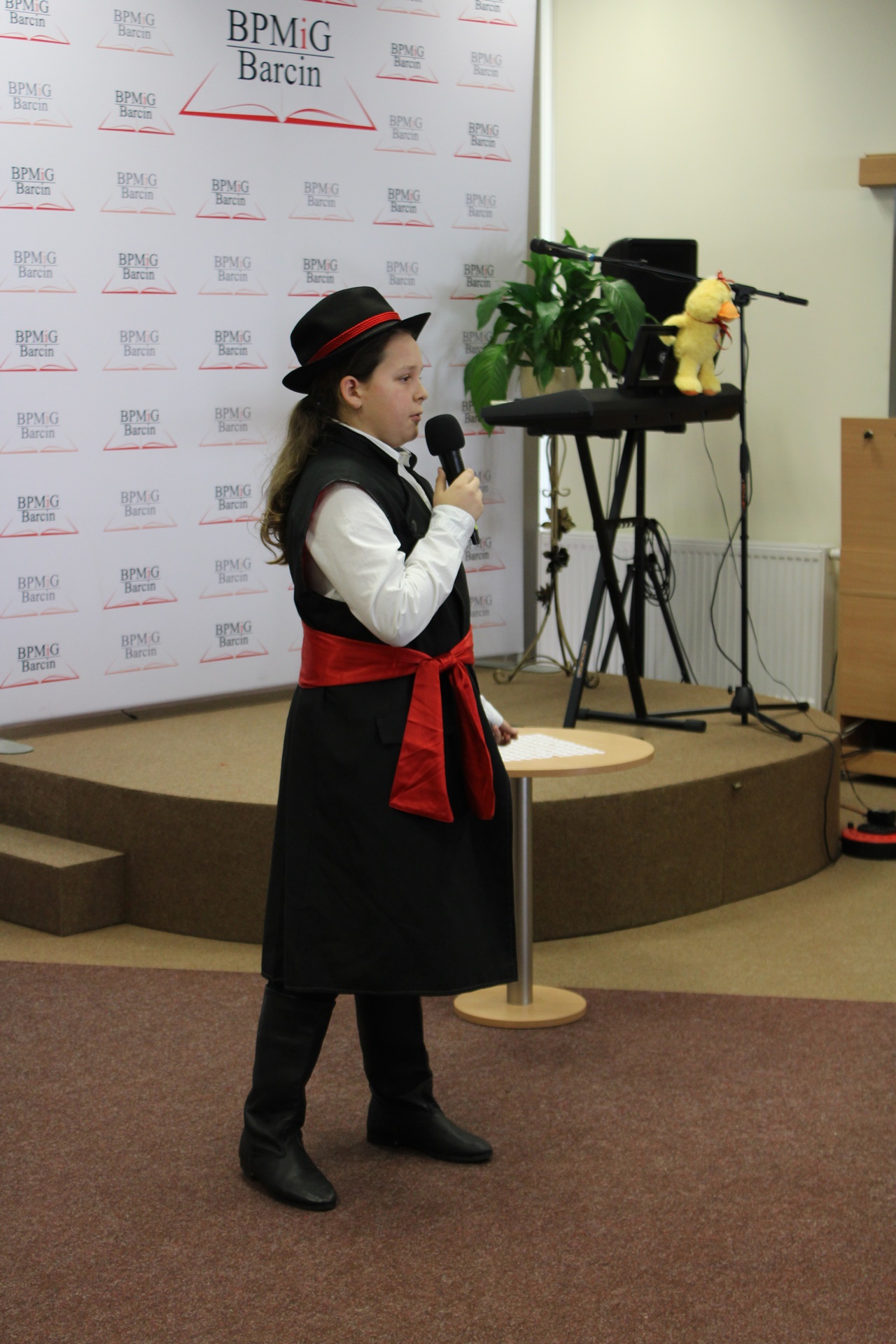 Chłopak z długimi włosami ubrany w czarny kapelusz obwiązany czerwoną wstążką, czarny kubrak bez rękawów przewiązany szeroką czerwoną szarfą, białą koszulę i czarne buty, występuje przed publicznością.