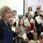Dyrektor Miejskiej i Powiatowej Biblioteki Publicznej w Żninie wypowiada się do mikrofonu, w tle stoją nagrodzeni uczestnicy i uczestniczki konkursu