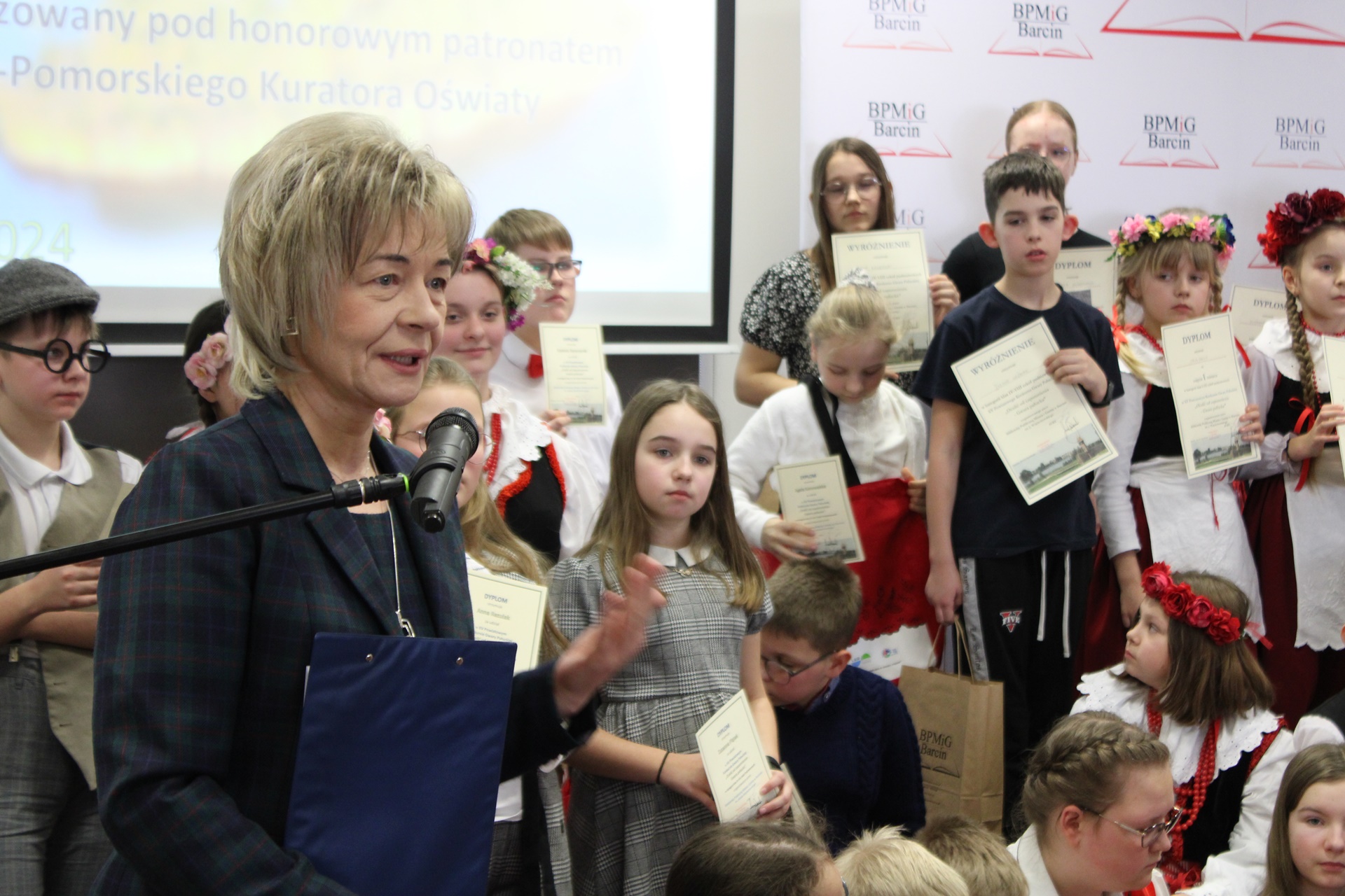 Dyrektor Miejskiej i Powiatowej Biblioteki Publicznej w Żninie wypowiada się do mikrofonu, w tle stoją nagrodzeni uczestnicy i uczestniczki konkursu