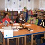6 nastolatek i jedna bibliotekarka, siedzą wokół stołów i trzymają otwarte przed sobą ksiażki