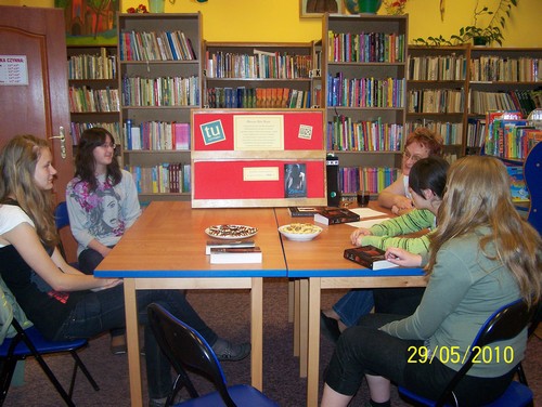 5 kobiet siedzi przy stołach i rozmawia ze sobą. Na stole stoi tablica do której przyczepiono okładkę omawianej książki oraz inne informacje dotyczące DKK