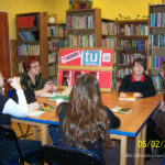 4 nastolatki i bibliotekarka prowadząca spotkanie siedzą przy stołach, przed nimi leżą książki, na stole stoi tablica na której przyczepione jest logo dkk, logo Instytutu Książki, grafika z napisem "TU CZYTAMY", oraz okładka omawianej ksiażki