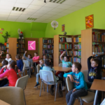 Dzieci razem z opiekunami siedzą przy stolikach pomiędzy stolikami chodzi bibliotekarka prowadząca zajęcia.