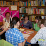 6 kobiet siedzących przy stołach jedna trzyma otwartą książkę inna czyta tekst z kartki A4
