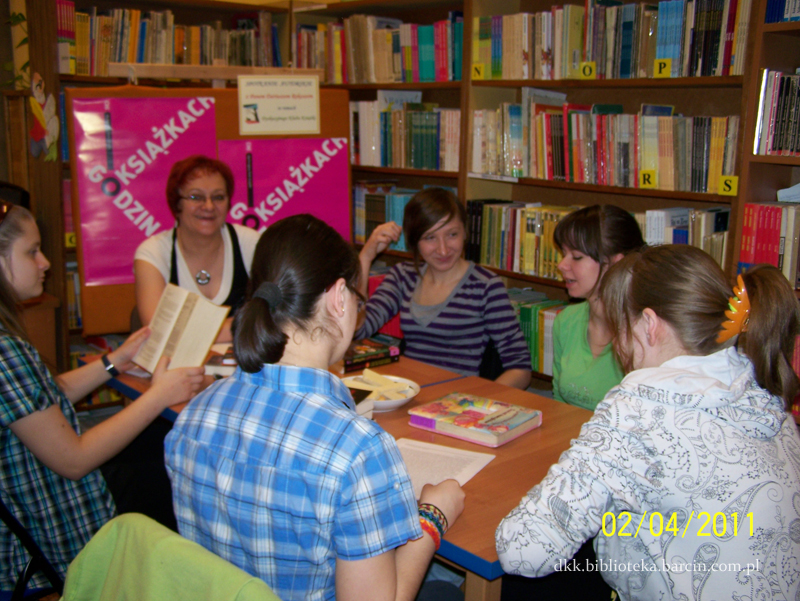 6 kobiet siedzących przy stołach jedna trzyma otwartą książkę inna czyta tekst z kartki A4