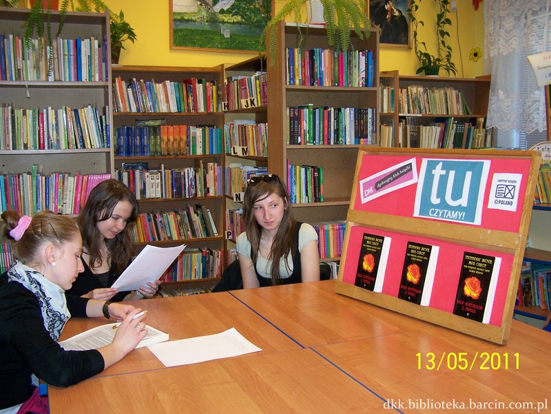 3 uczestniczki spotkania, dwie czytaja coś z kartek papieru trzecia przygląda sie tablicy z logo DKK, instytutu ksiażki hasłem "tu czytamy" i dwiema omawianymi ksiażkami