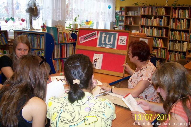 cztery uczestniczki i prowadząca siedzą wspólnie przy stołach, dwie uczestniczki czytają książki, prowadząca pokazuje tablicę dotyczącą spotkania DKK 